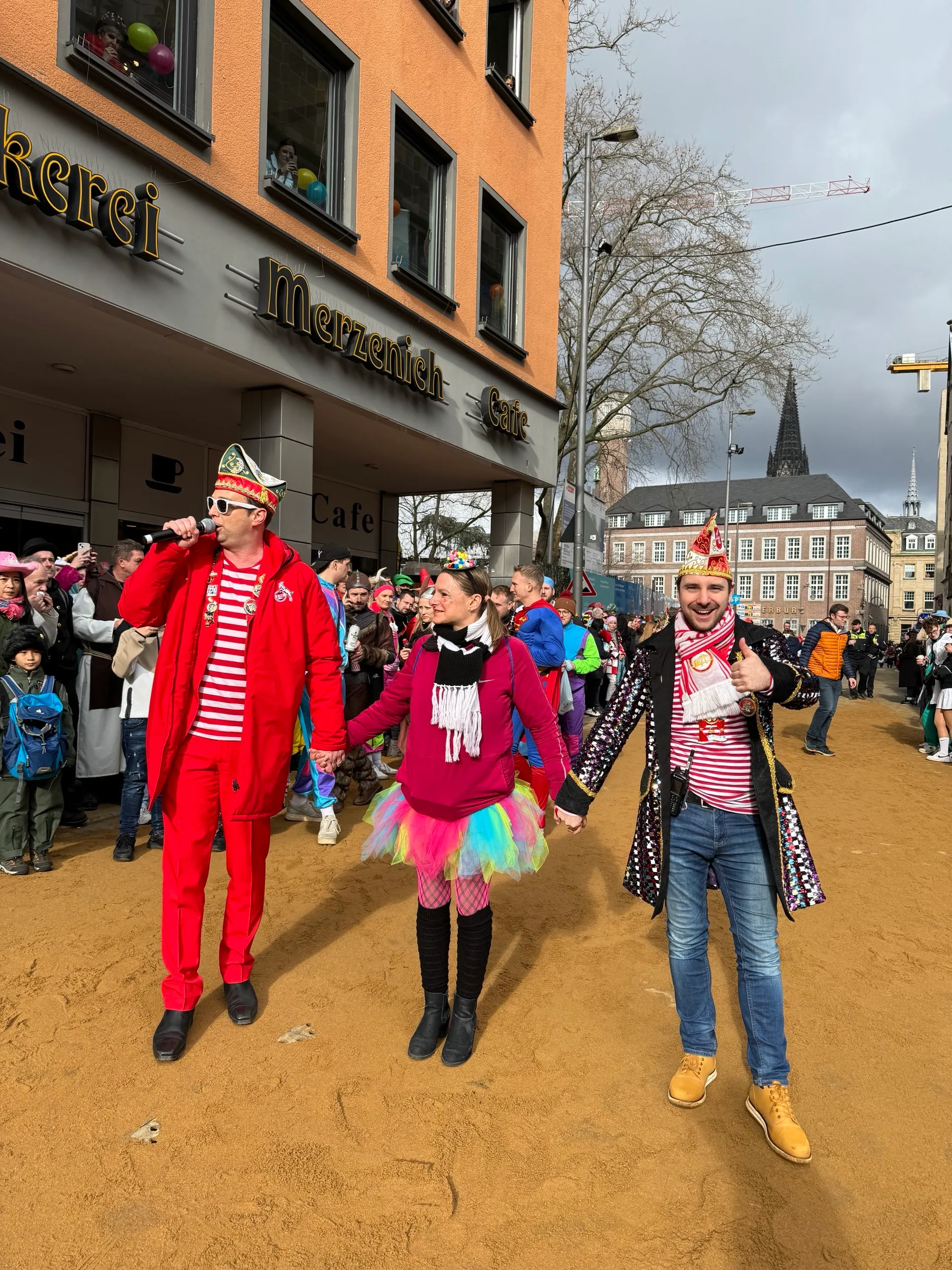 Rosenmontagszug 2026 vor der Bäckerei Merzenich: Moderator Lukas Wachten (Dorint-Tribüne) mit Mikrofon auf der Sandbahn, Domspitzen im Hintergrund